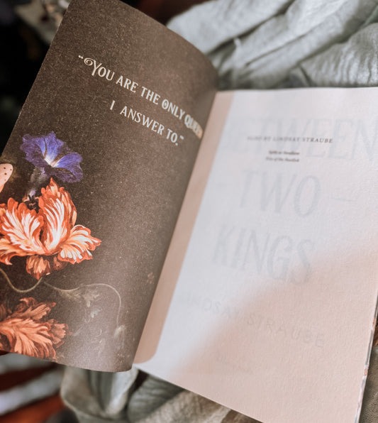 Book titled 'Between Two Kings' by Lindsay Straube on a textured surface with a candle and flowers.