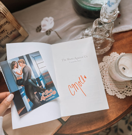 Book titled 'The Shots Against Us' by Cassandra Moll on a wooden surface with a doily and glassware in the background.