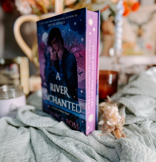 Book titled 'A River Enchanted' by Rebecca Ross on a soft surface with candles and a cup.