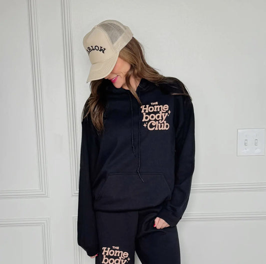 Person wearing a black hoodie and pants with 'Homebody Club' text in a forest setting