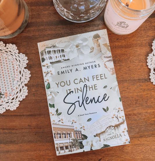 Book titled 'You Can Feel It in the Silence' by Emily A. Myers on a wooden surface with a glass of orange juice and a candle.
