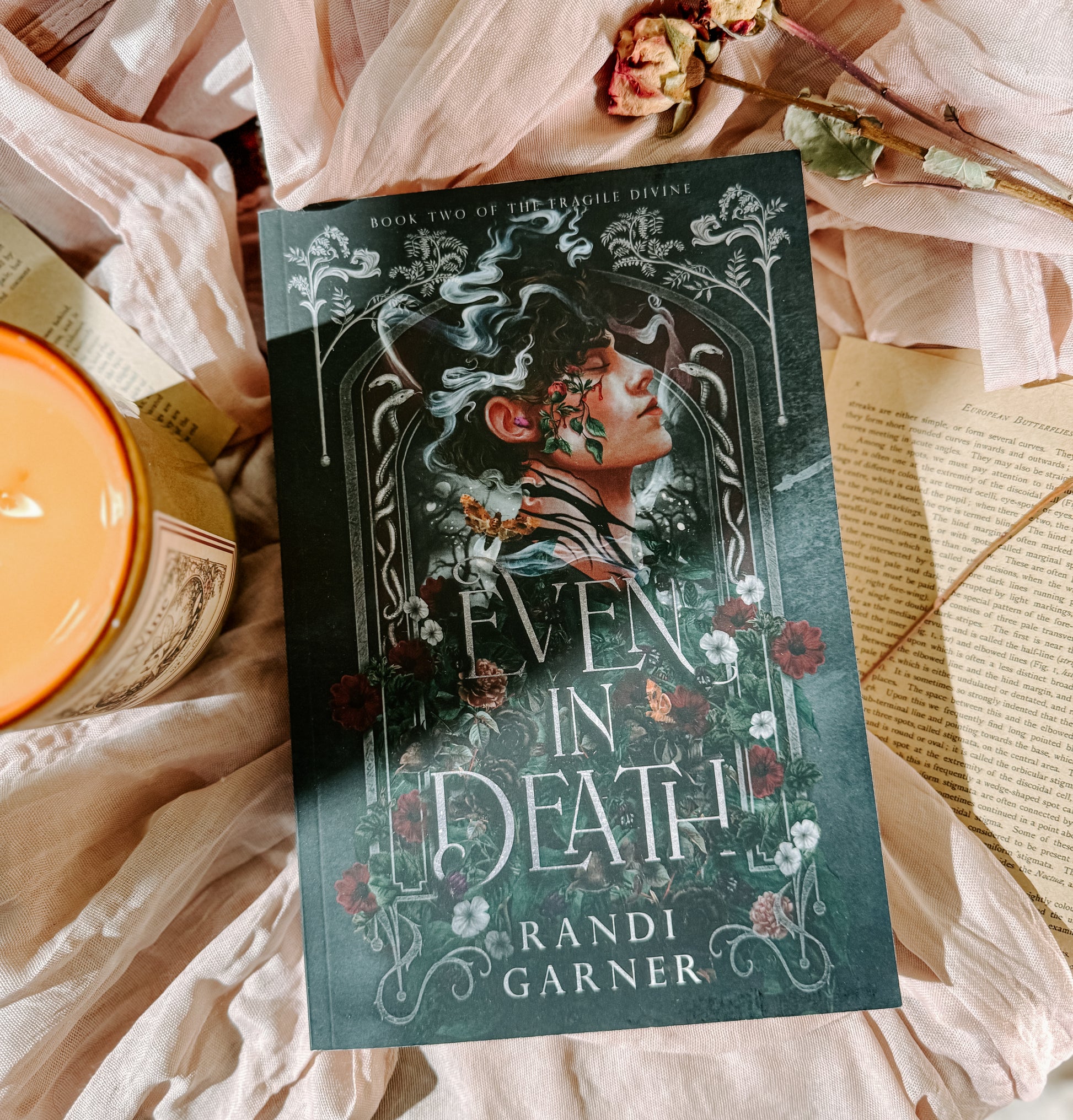 Book titled 'Even in Death' by Randi Garner on a soft fabric background with flowers and a candle.