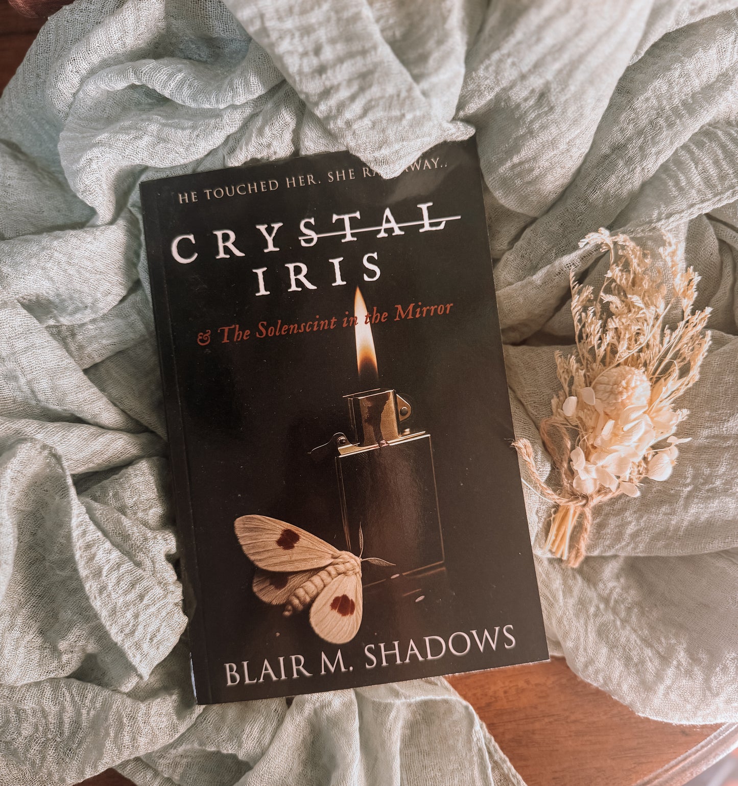 Book titled 'Crystal Iris' by Blair M. Shadows on a textured surface with a small bouquet of flowers.