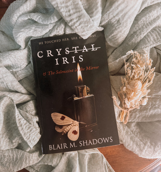 Book titled 'Crystal Iris' by Blair M. Shadows on a textured surface with a small bouquet of flowers.