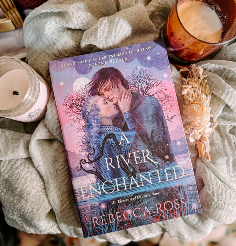 Book titled 'A River Enchanted' by Rebecca Ross on a soft surface with candles and a cup.
