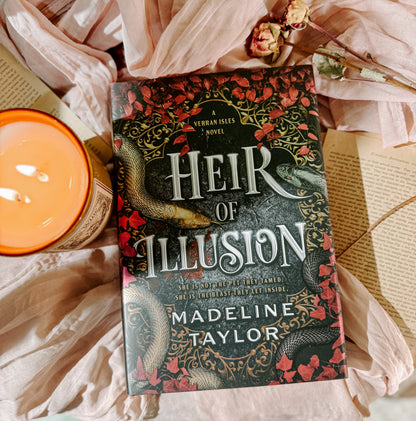 Book titled 'Heir of Illusion' by Madeline Taylor on a soft fabric background with a candle and flowers.