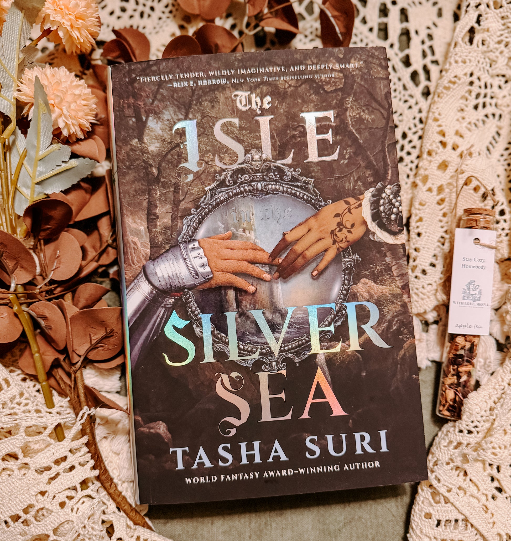 Book titled 'The Isle of Silver Sea' by Tasha Suri on a decorative background with flowers and lace.