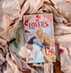Book titled 'The Lovers' by Kristin Cast on a soft fabric background