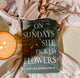 Book titled 'On Sundays She Picked Flowers' by Yah Yah Scholfield on a soft pink fabric background with a candle and dried flowers.