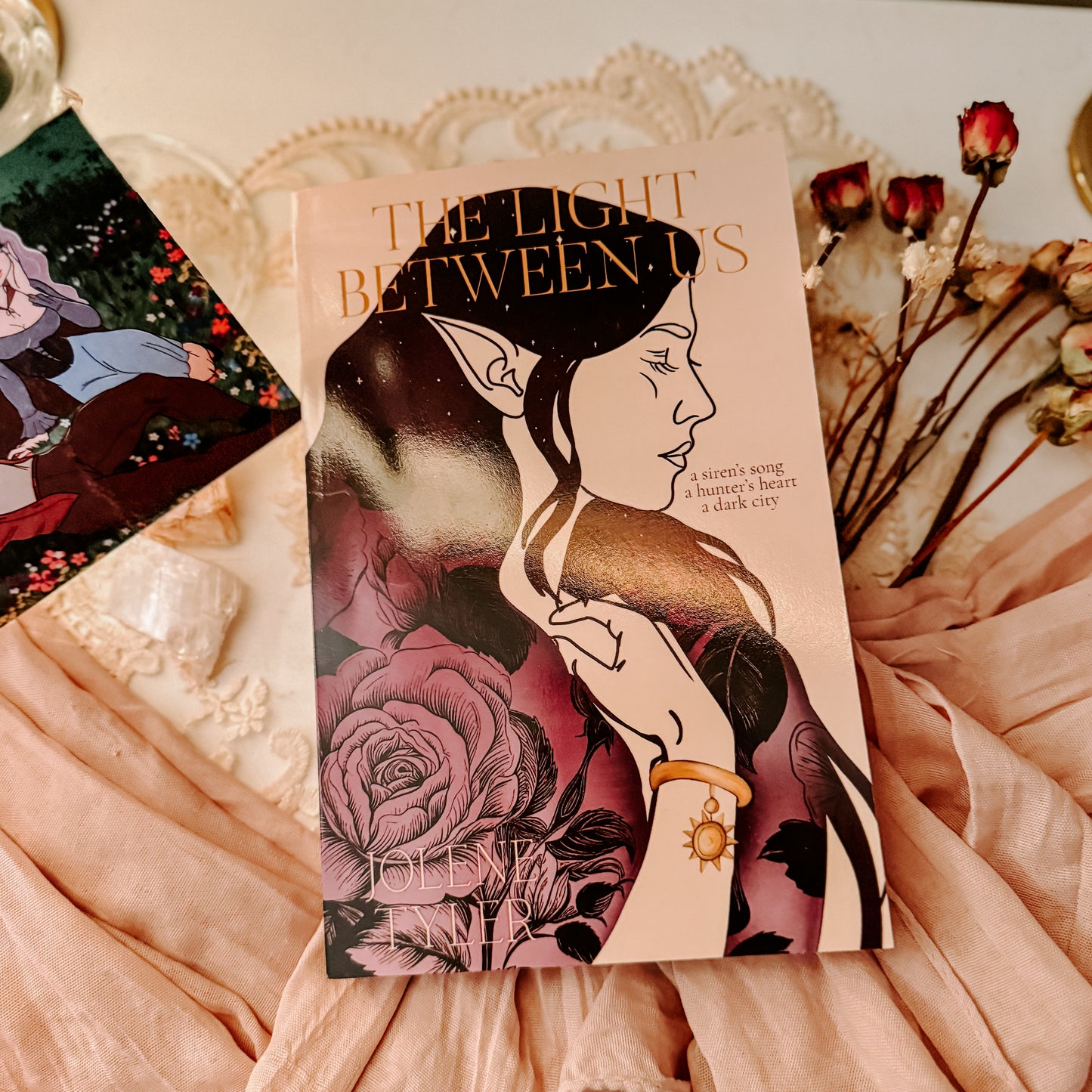 Book titled 'The Light Between' with fantasy-themed cover featuring two faces and floral elements, placed on a soft fabric background.