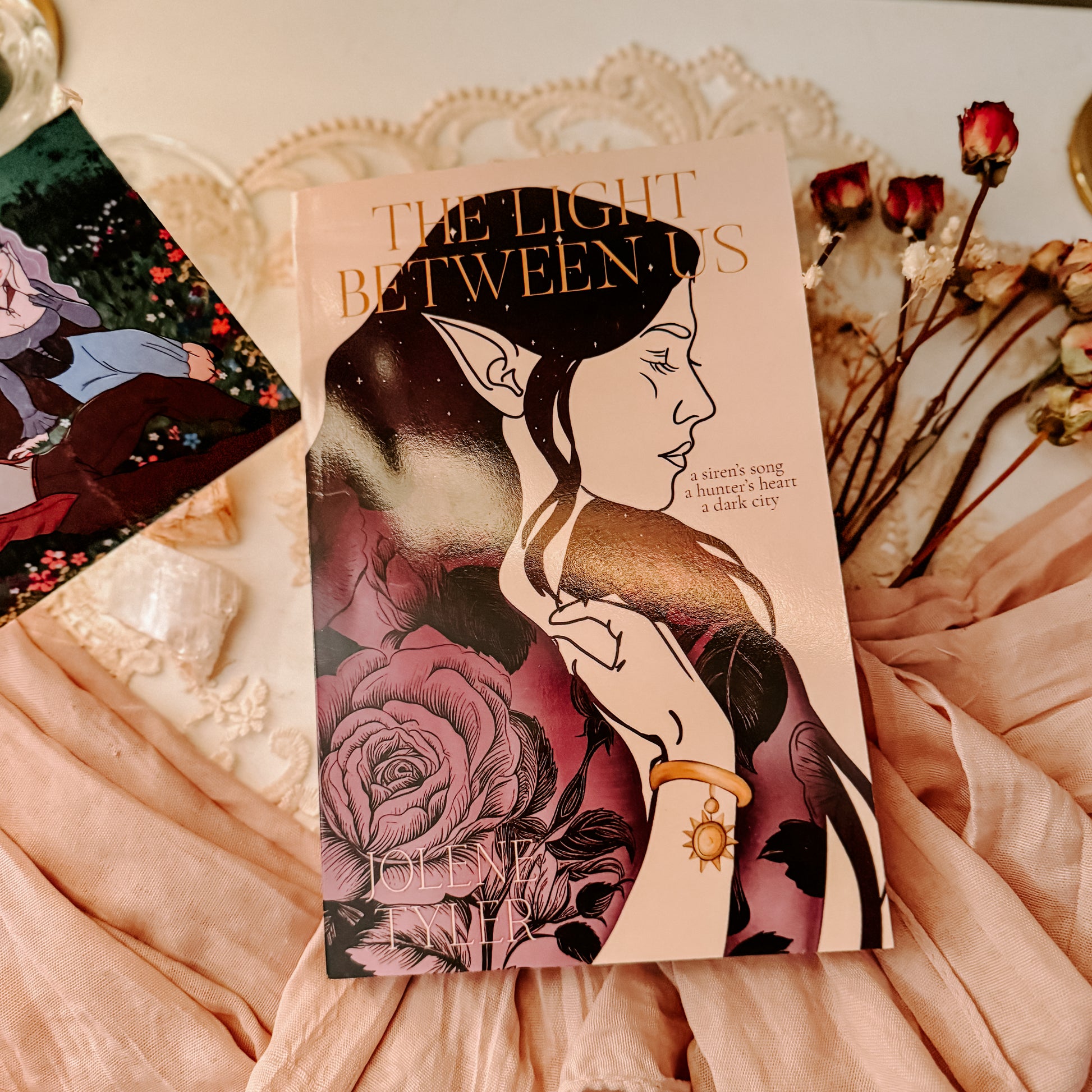 Book titled 'The Light Between' with fantasy-themed cover featuring two faces and floral elements, placed on a soft fabric background.