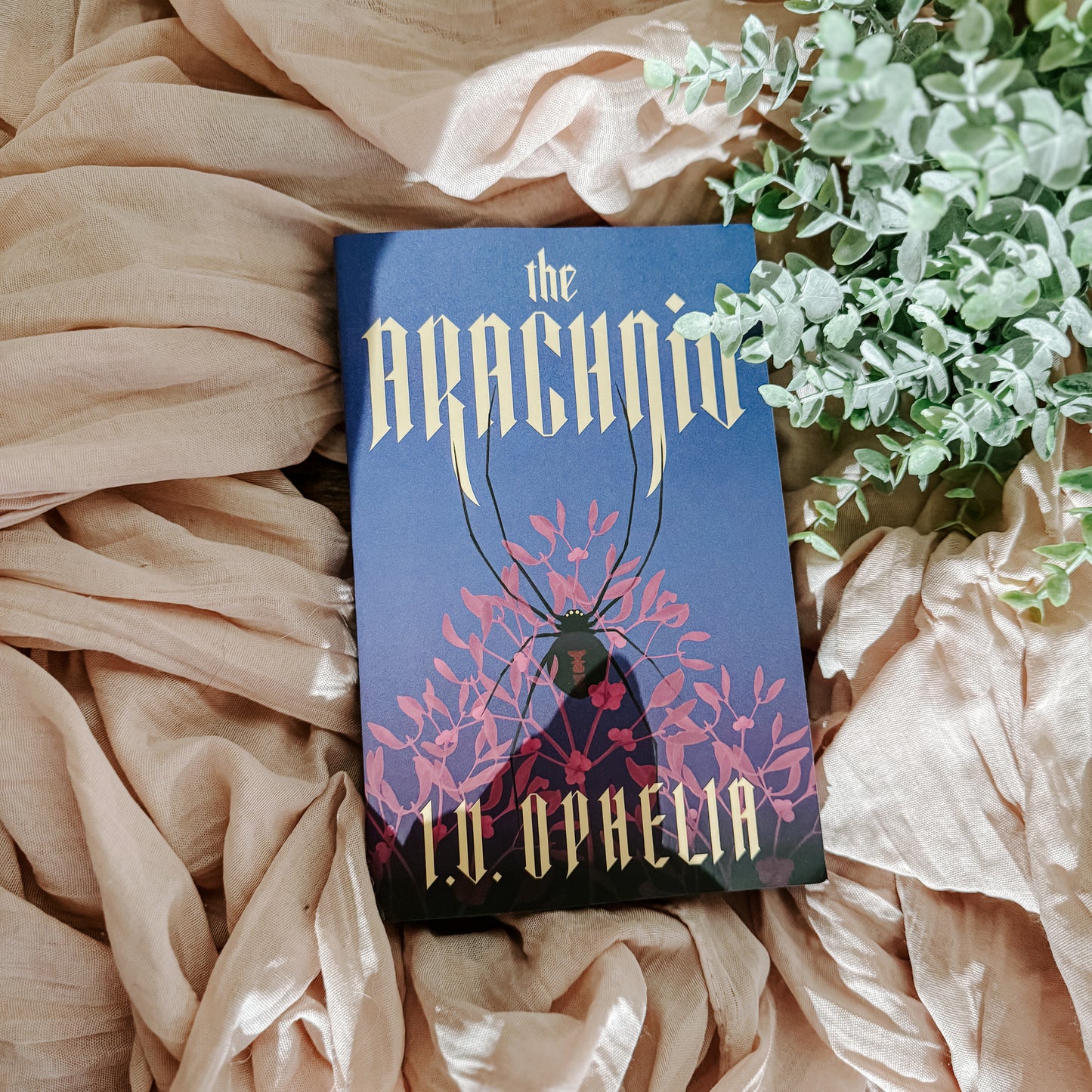 Book titled 'The Arachnid' by I.V Ophelia on a soft fabric background with greenery.