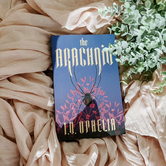 Book titled 'The Arachnid' by I.V Ophelia on a soft fabric background with greenery.
