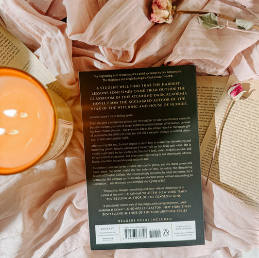 Book titled 'An Academy for Liars' by Alexis Henderson on a soft pink fabric background with a candle and flowers.