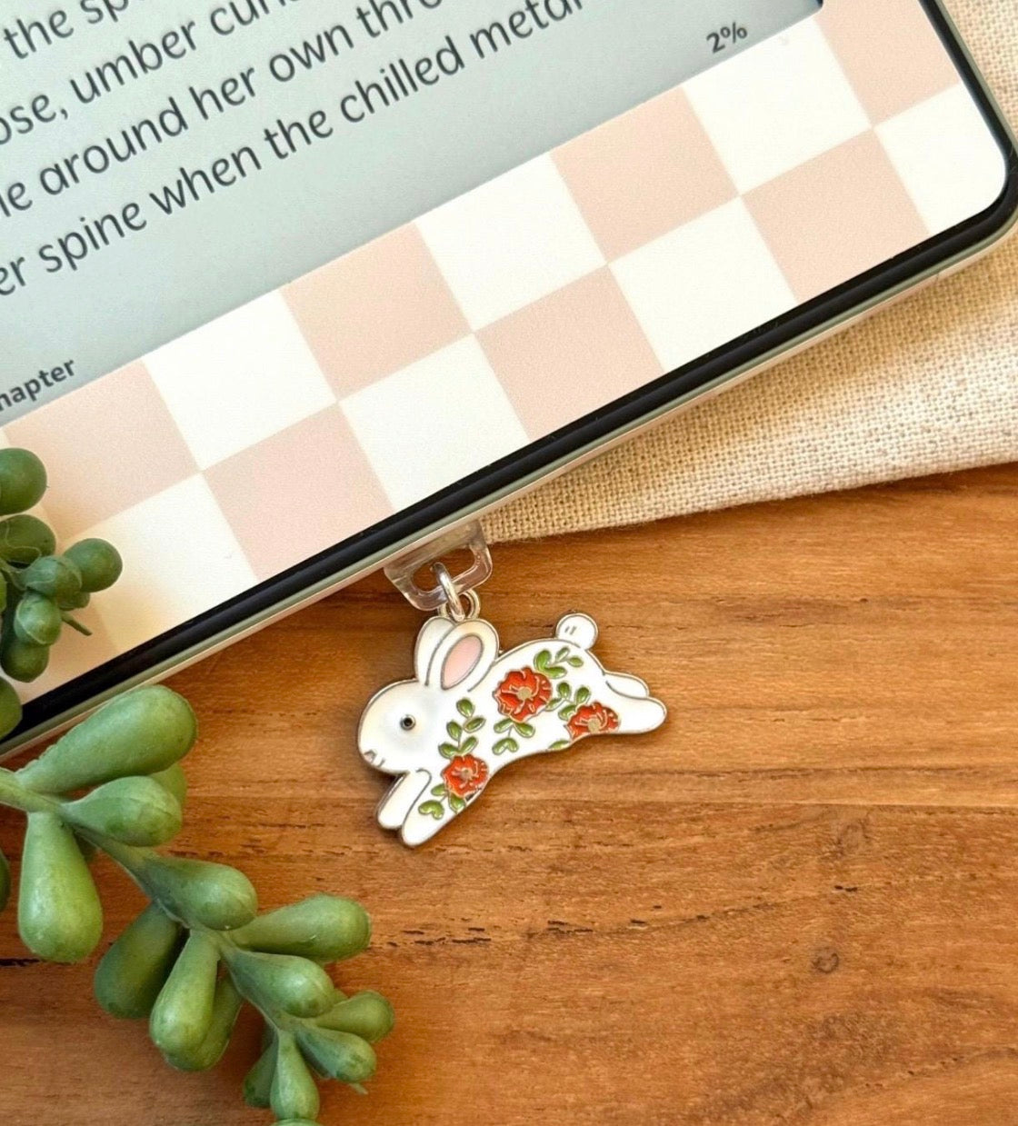 Silver rabbit charm with floral design on a wooden surface next to a smartphone displaying a book.