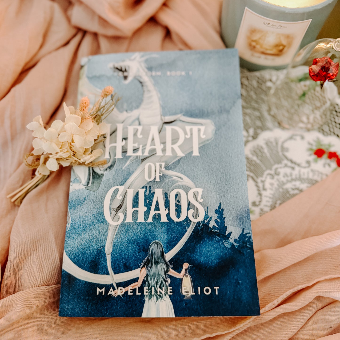 Book titled 'Heart of Chaos' by Madeleine Eliot on a soft fabric background with a candle and flowers.