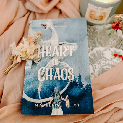 Book titled 'Heart of Chaos' by Madeleine Eliot on a soft fabric background with a candle and flowers.