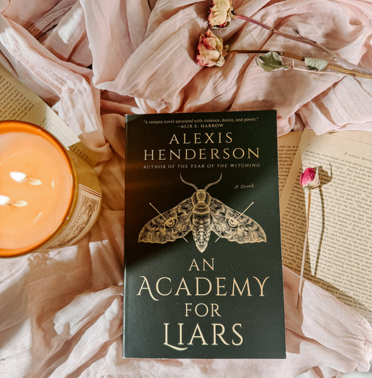 Book titled 'An Academy for Liars' by Alexis Henderson on a soft pink fabric background with a candle and flowers.