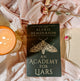 Book titled 'An Academy for Liars' by Alexis Henderson on a soft pink fabric background with a candle and flowers.
