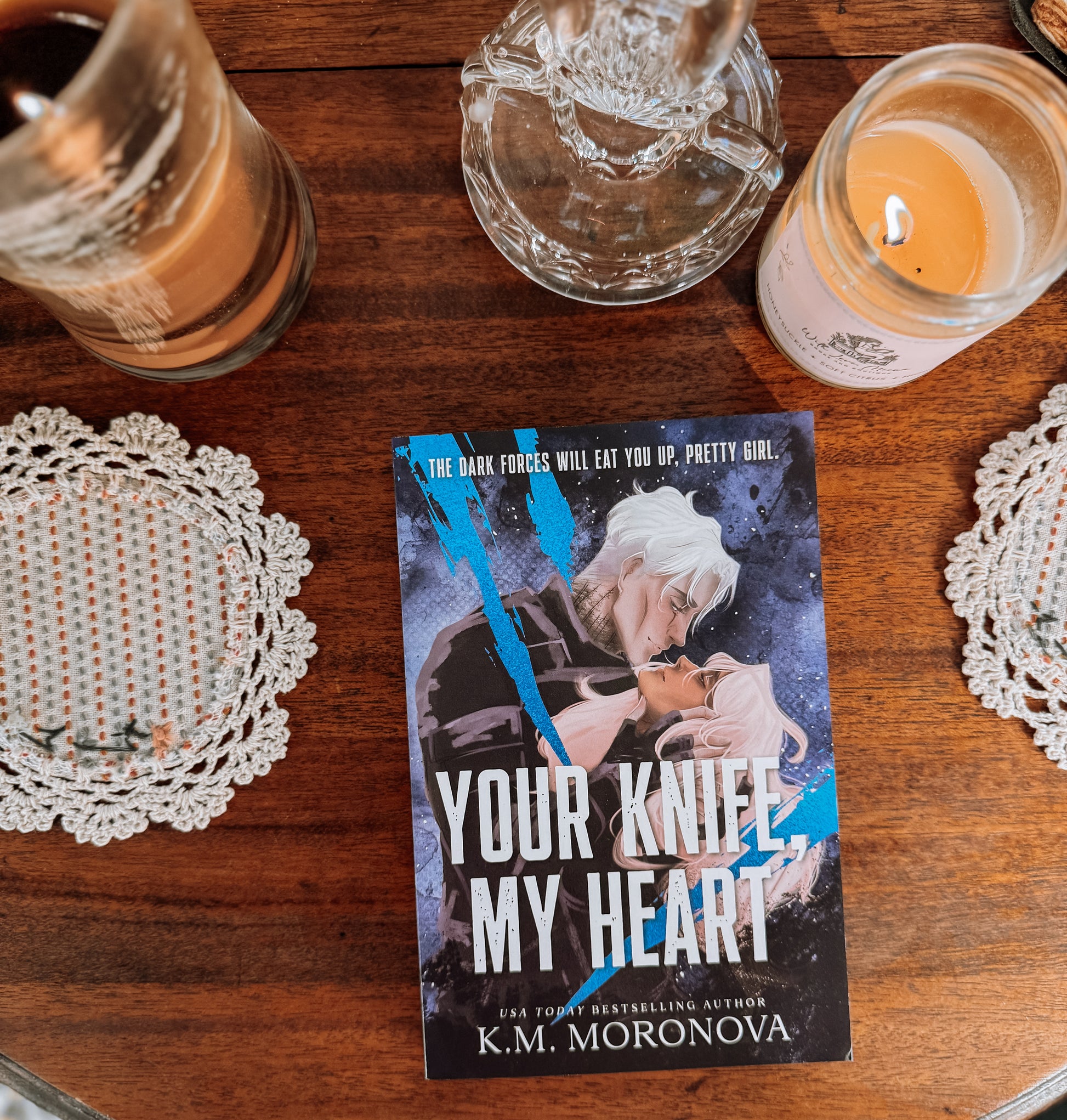 Book titled 'Your Knife My Heart' by K.M. Moronova on a wooden surface with candles and doilies.