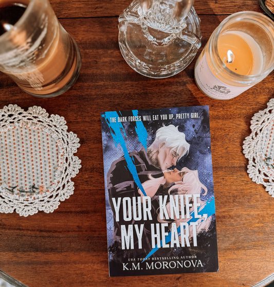 Book titled 'Your Knife My Heart' by K.M. Moronova on a wooden surface with candles and doilies.