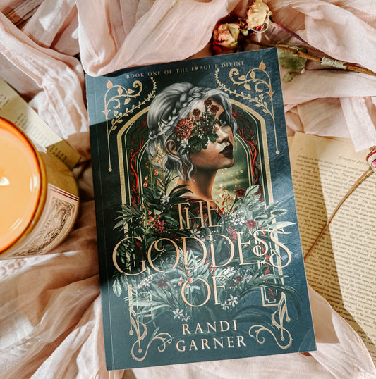Book titled 'The Goddess of' by Randi Garner on a soft fabric background with a candle and flowers.