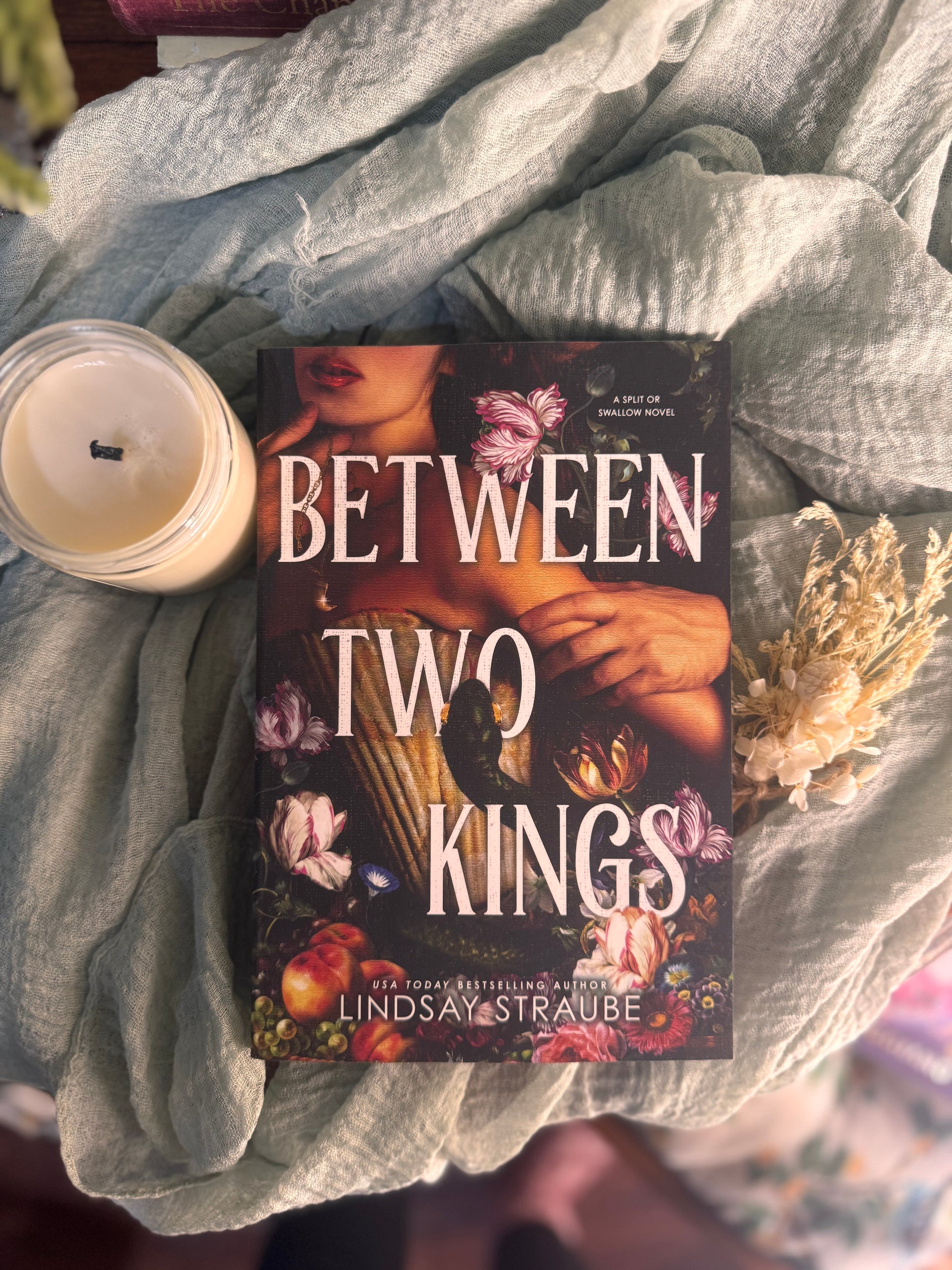Book titled 'Between Two Kings' by Lindsay Straube on a textured surface with a candle and flowers.