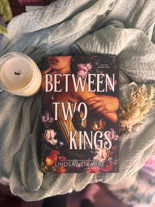 Book titled 'Between Two Kings' by Lindsay Straube on a textured surface with a candle and flowers.