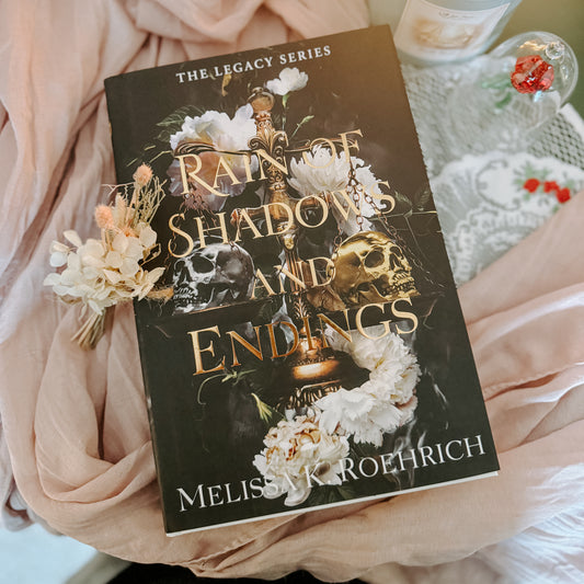 Book titled 'Rain of Shadows and Endings' from The Legacy Series held by a person with flowers.