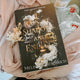 Book titled 'Rain of Shadows and Endings' from The Legacy Series held by a person with flowers.