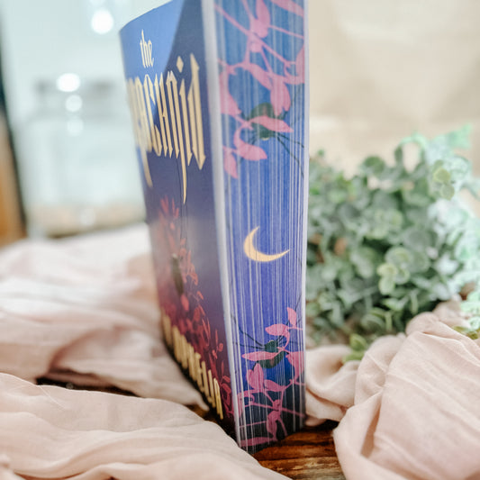 Book titled 'The Arachnid' by I.V Ophelia on a soft fabric background with greenery.