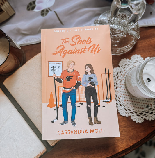 Book titled 'The Shots Against Us' by Cassandra Moll on a wooden surface with a doily and glassware in the background.