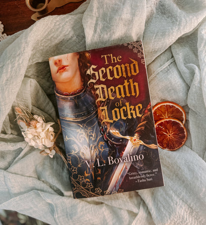 Book titled 'The Second Death of Locke' by L. Bovalino on a textured fabric background with dried oranges and flowers.