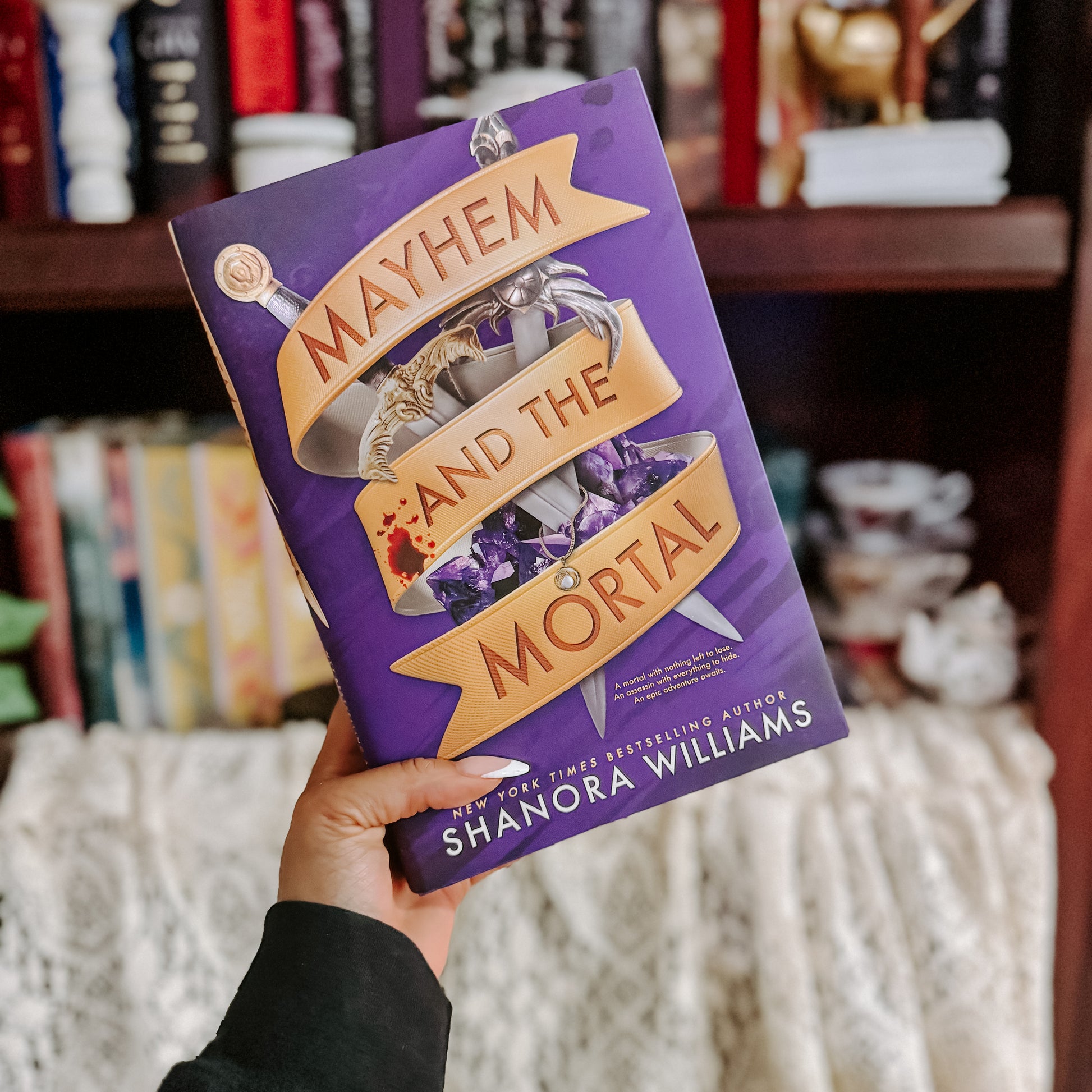 Book titled 'Mayhem and the Mortal' by Shanora Williams held in front of a bookshelf.