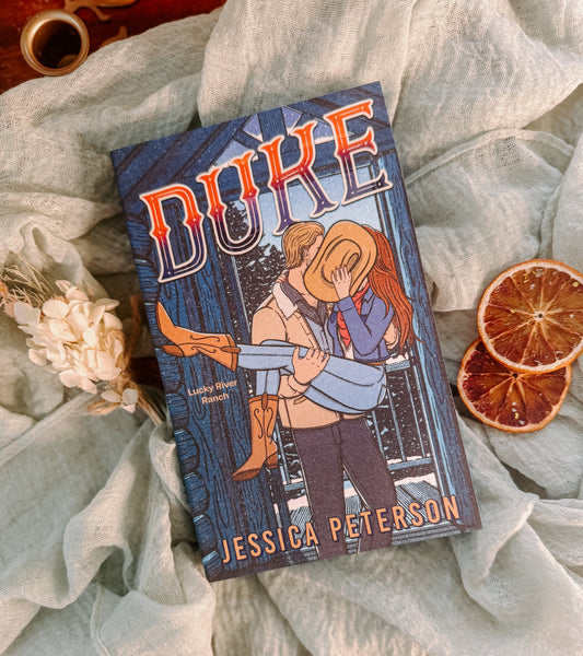 Book titled 'Duke' by Jessica Peterson on a textured surface with dried flowers and oranges.