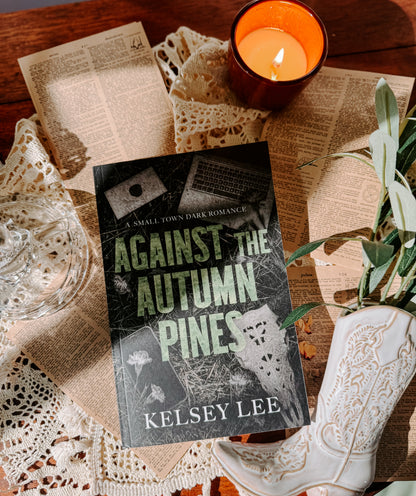 Book titled 'Against the Autumn Pines' by Kelsey Lee on a wooden surface with a candle and lace fabric.