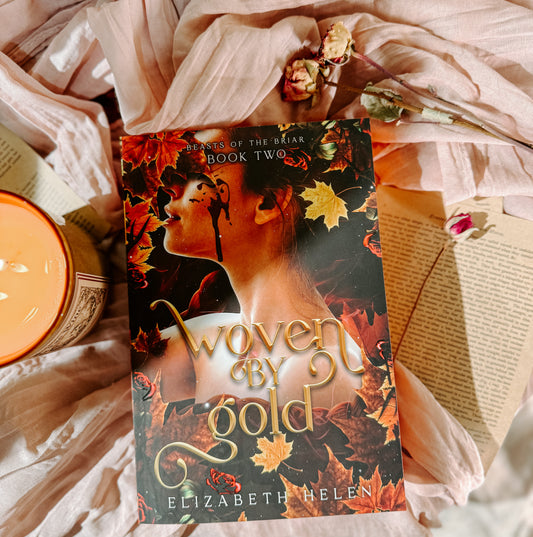Book titled 'Woven by Gold' by Elizabeth Helen on a soft fabric background with a candle.