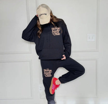 Person wearing navy 'Homebody Club' hoodie and sweatpants with a beige cap, standing against a white paneled wall.